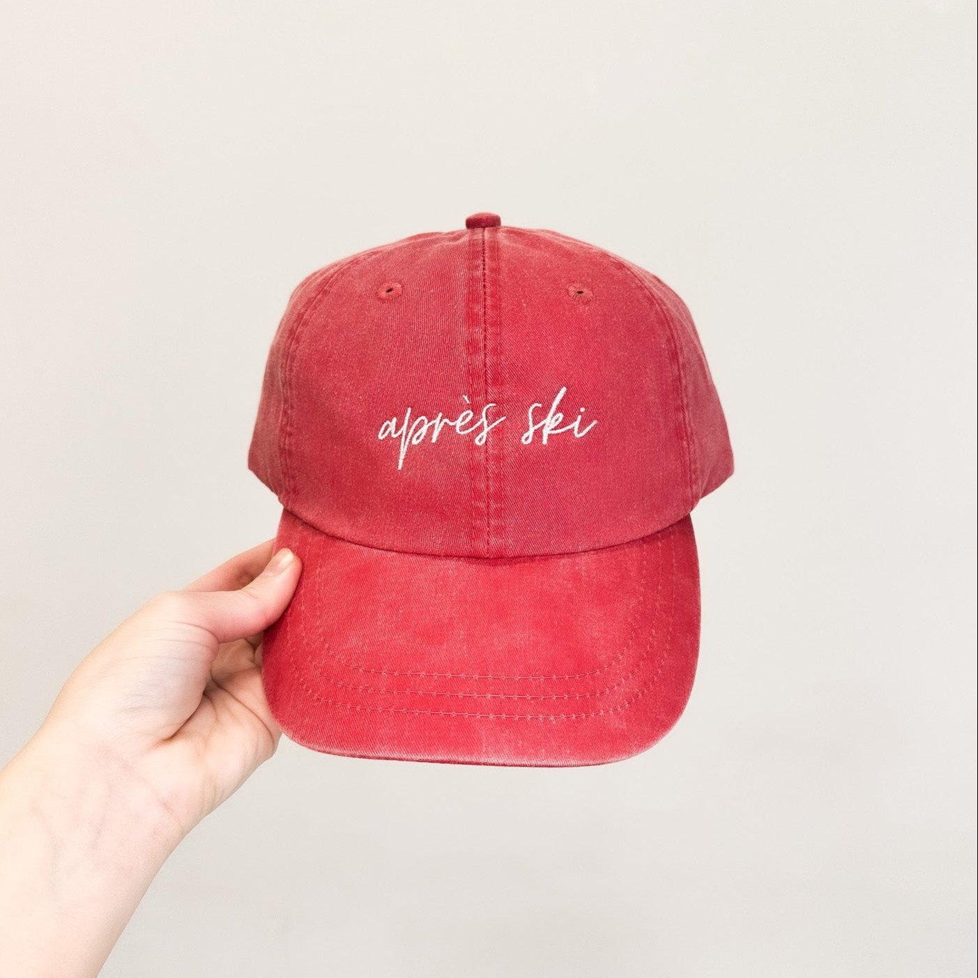 A hand holding a red baseball cap with 'Aprés Ski' embroidered in white on the front.